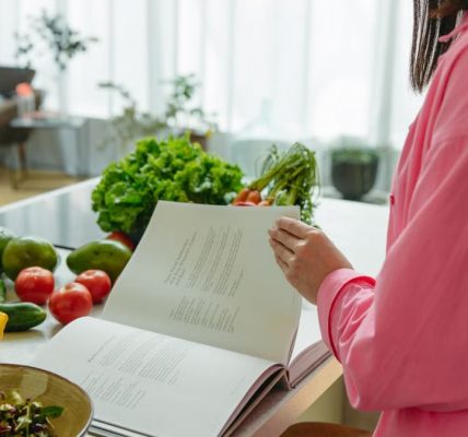 A woman in a kitchen flipping through a cookbook surrounded by fresh vegetables.