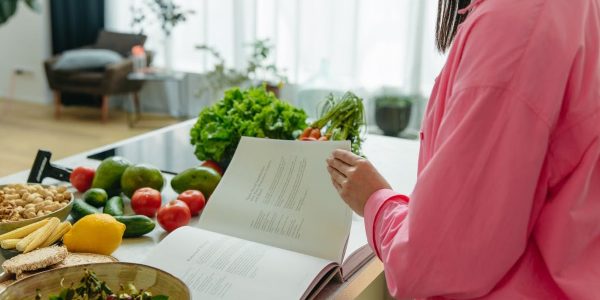 A woman in a kitchen flipping through a cookbook surrounded by fresh vegetables.